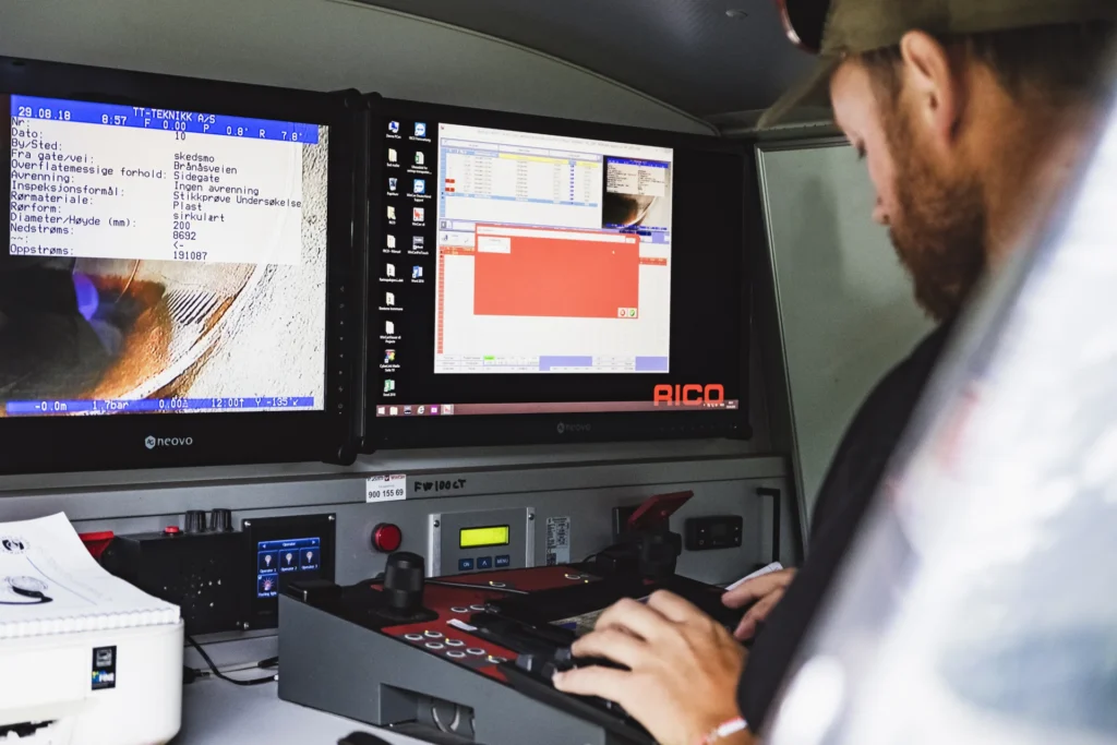 En person betjener et kontrollpanel inne i en spesialbil, konsentrert om to skjermer som viser programvaregrensesnitt og data. Oppsettet inkluderer en rekke knapper og kontroller, med elektronisk utstyr strategisk plassert i nærheten.