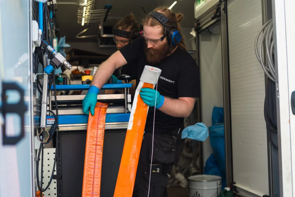 I et utstyrsfylt verksted, der Rørfornying i Solrabben borettslag er i gang, tar en person med skjegg og briller på seg beskyttende øreklokker og hansker for å inspisere lange oransje strimler, mens en annen person er delvis synlig i bakgrunnen.