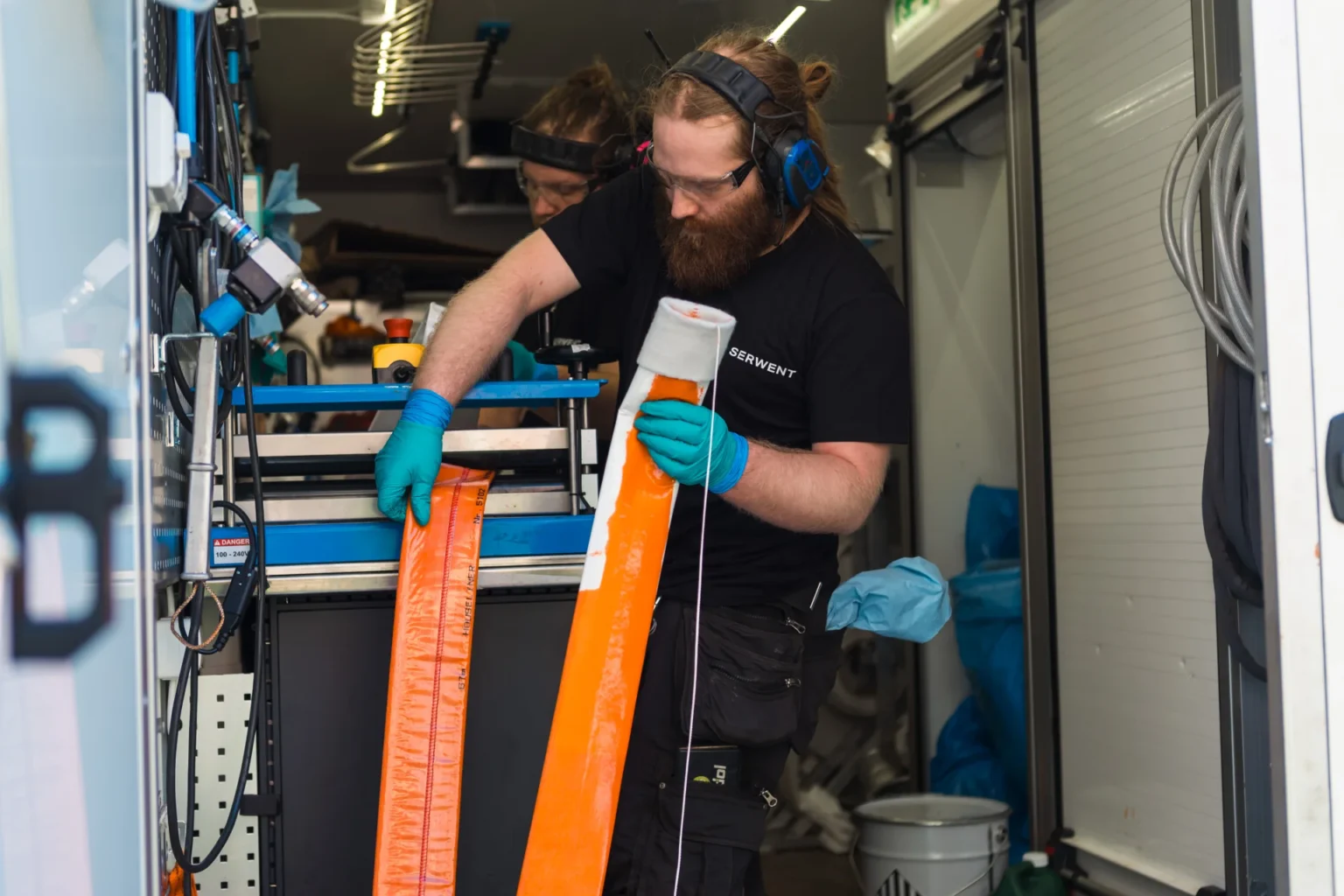 I et utstyrsfylt verksted, der Rørfornying i Solrabben borettslag er i gang, tar en person med skjegg og briller på seg beskyttende øreklokker og hansker for å inspisere lange oransje strimler, mens en annen person er delvis synlig i bakgrunnen.