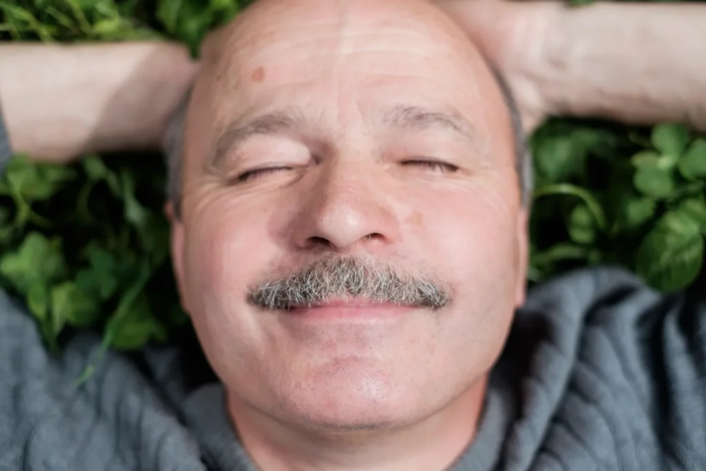 Movember Foundation En person med bart, som deltar i Movember, ligger på en seng med grønne blader. Øynene deres er lukket, og et fornøyd smil pryder ansiktet deres. De hviler armene bak hodet mens de har på seg en grå genser.