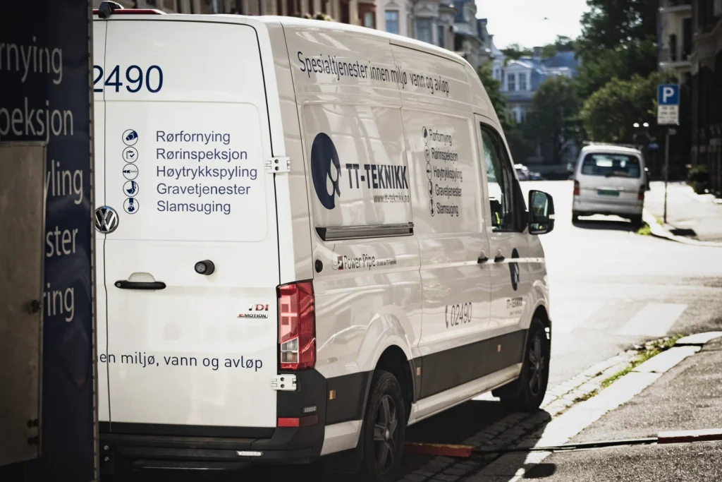 En hvit bruksbil med firmamerke og kontaktinformasjon fra Omsorgsbygg Oslo KF står parkert i en bygate. Teksten, på norsk, viser tjenester som rørinspeksjon og rydding. Sollys kaster skygger på bygninger og trær i nærheten.