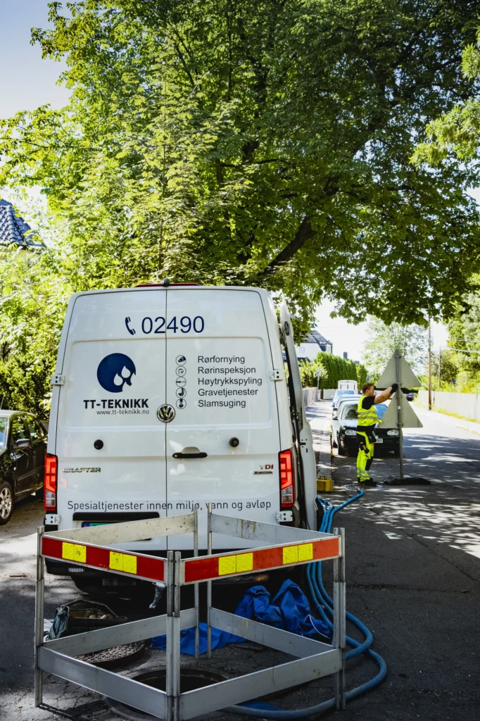 En varebil med «TT-Teknikk» og telefonnummer står parkert i en gate i Sinsenbyen. En arbeider i lyst antrekk står i nærheten og betjener utstyr koblet til varebilen. Trær og et annet kjøretøy er synlige i bakgrunnen på en solrik dag.