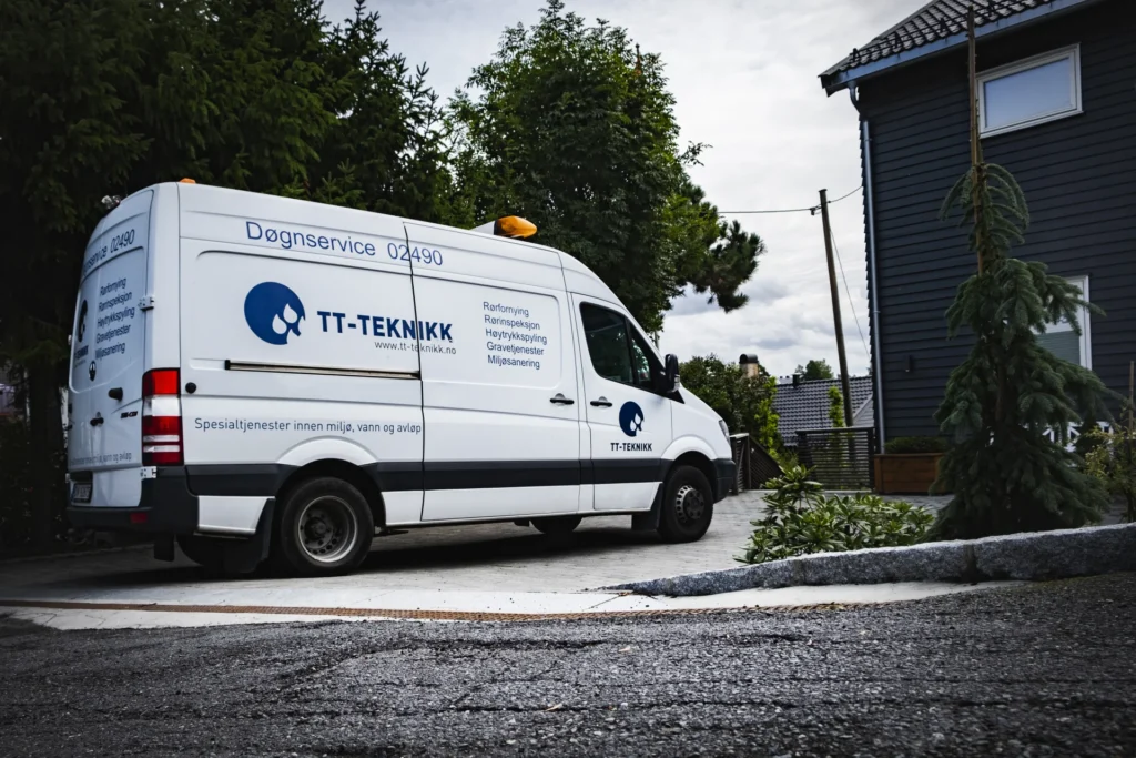 07-ror-inspeksjon-9 En hvit varebil med «TT-TEKNIKK» og tjenester som «fornye rør» oppført på siden står parkert i en innkjørsel ved siden av et moderne grått hus. Et lite eviggrønt tre og frodige grøntområder gir et rolig bakteppe til scenen.