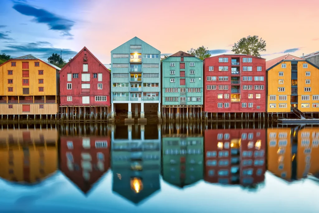 Fargerike trebygninger i Trondheim står ved en rolig elv, og reflekterer levende nyanser av rødt, blått, grønt og gult i det stille vannet. En rosa og blå solnedgangshimmel forsterker denne pittoreske norske scenen.