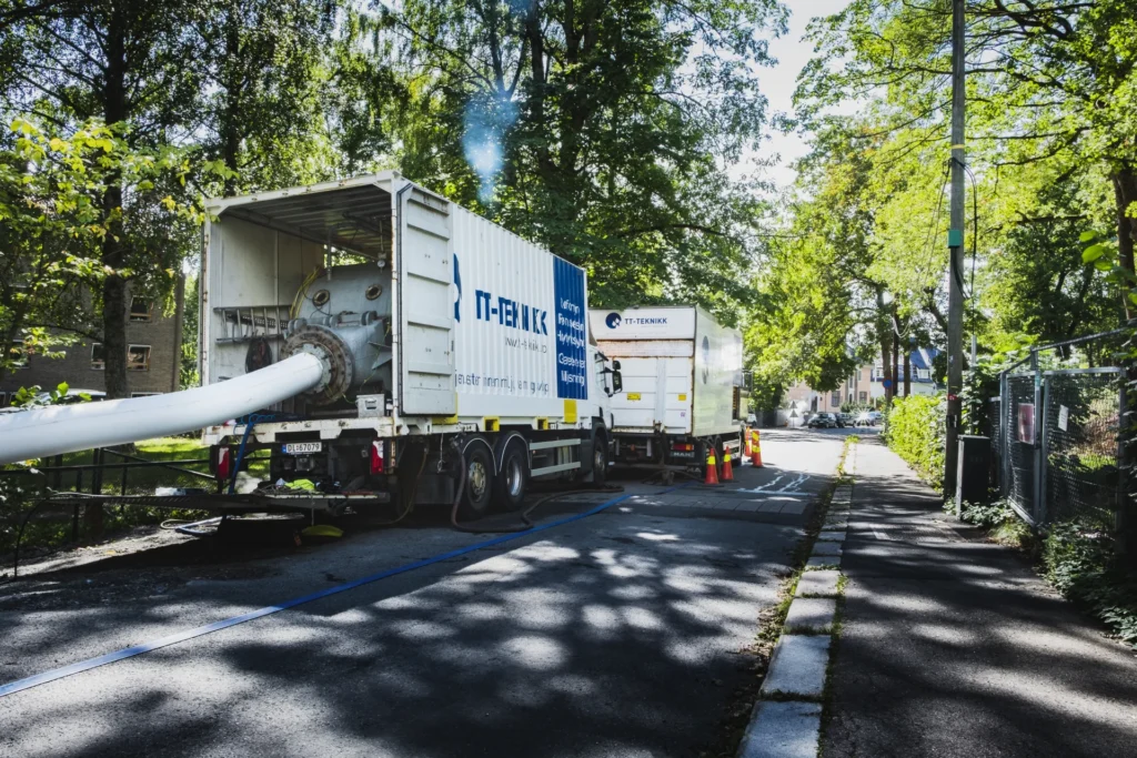 To store lastebiler med påmontert ISO-sertifiseringsutstyr parkert på en smal vei omgitt av trær. En hvit slange strekker seg fra en lastebil, og indikerer industriarbeid. Sterkt sollys siler gjennom trærne og kaster skygger på bakken.