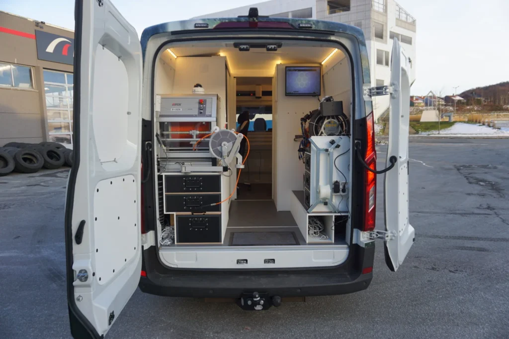 Bærekraftig rørinspeksjon Åpen rørinspeksjonsbil med teknisk utstyr inne, parkert utendørs. Utstyret inkluderer skjermer, kabler og oppbevaringsskuffer, noe som foreslår et mobilt laboratorium eller servicekjøretøy. I nærheten ligger bygninger og en stabel med dekk under overskyet himmel.