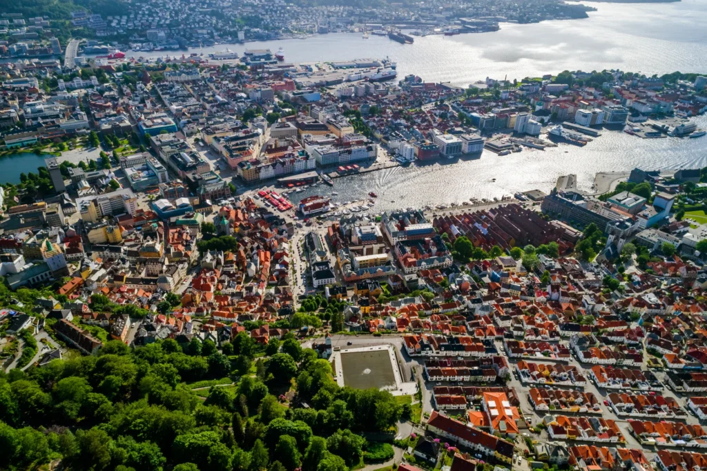 Luftfoto av en kystby som minner om Bergen, med en livlig havn. Bildet viser tettpakkede bygninger med røde tegltak, svingete gater og frodige grønne områder. I bakgrunnen er en vannmasse synlig, oversådd med båter og brygger langs kysten.
