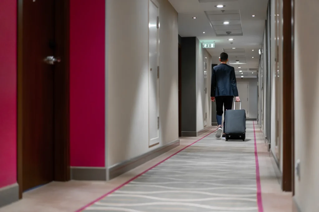 Vi utfører rens av ventilasjon for alle hoteller! En person i dress går ned en hotellgang og trekker en koffert. Korridoren har et mønstret teppe og rosa aksentvegger. Innfelt belysning og godt vedlikeholdt ventilasjon sørger for at plassen føles frisk, og leder dem mot det fjerne utgangsskiltet.