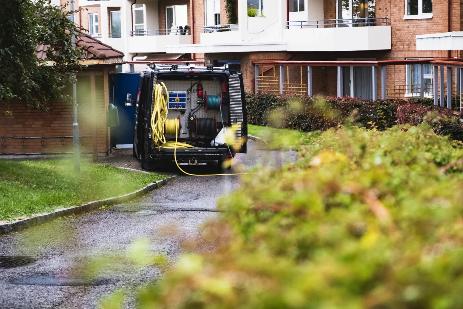 En bruksbil med utstyr og slanger, merket «Tette rør», står parkert i en boliggate, delvis skjult av en busk. I bakgrunnen ruver en bygård med balkonger, mens en litt uskarp hekk rammer inn forgrunnen.