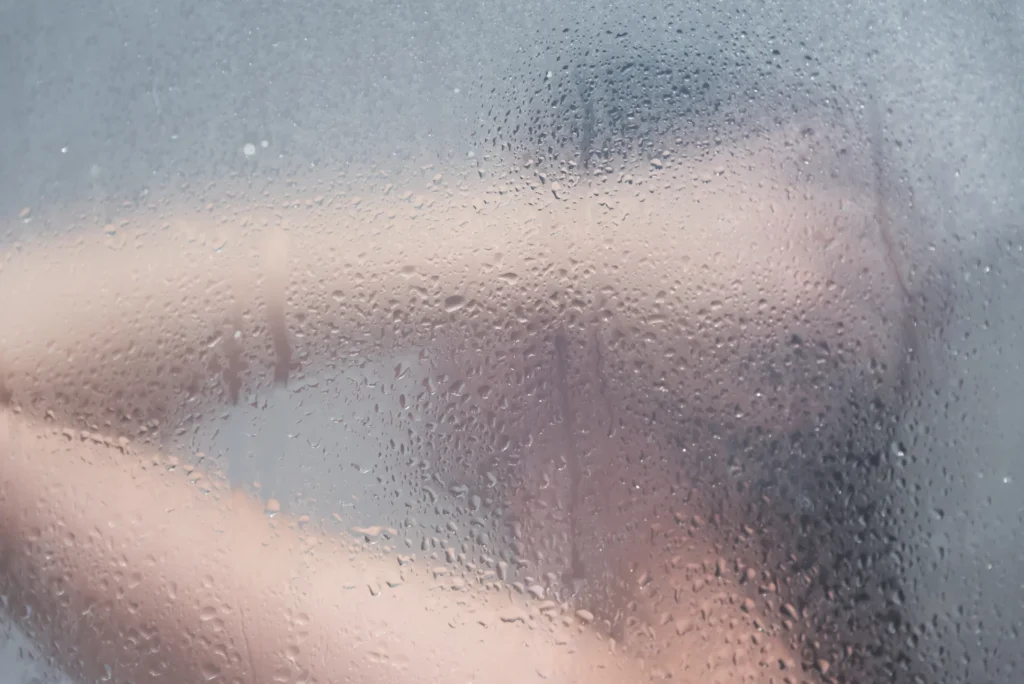 Hvilken vifte er best på badet? En uskarp person sees gjennom en tåkete, vannstripet glassflate, kanskje på et bad med vifte på badet som skaper virvler av kondens. Armene deres er litt hevet, med utydelige trekk, som formidler en følelse av mystikk og mykhet.
