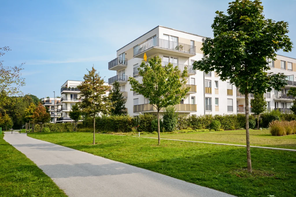Moderne bygårder med hvite fasader og balkonger, utstyrt med topp moderne ventilasjon i næringsbygg, er satt opp mot en klarblå himmel. En asfaltert sti vever seg gjennom en frodig grønn plen og trær i forgrunnen, og skaper et rolig bymiljø.