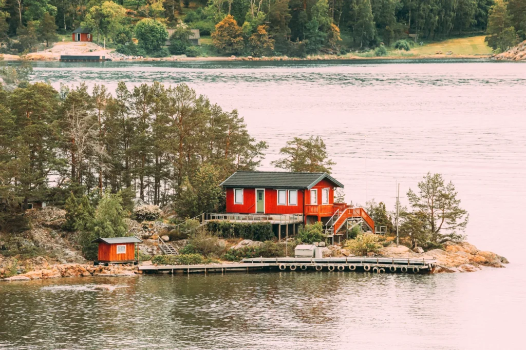 naturlig ventilasjon En rød hytte i skandinavisk stil med naturlig ventilasjon ligger på en liten steinete øy omgitt av stille vann. Øya er strødd med trær og et lite skur, mens i bakgrunnen er en skogkledd strandlinje synlig under en overskyet himmel.