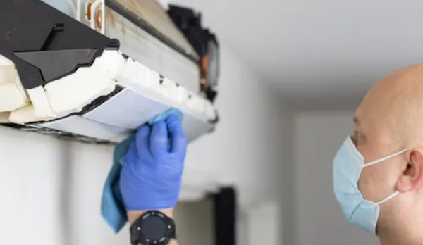 En person som har på seg en maske og blå hansker, rengjør flittig interiøret i en klimaanlegg med en klut, for å sikre optimalt vedlikehold. Vurderer ventilasjonsanlegg på arbeidsplass? Det er avgjørende for å opprettholde luftkvaliteten på arbeidsplassen.