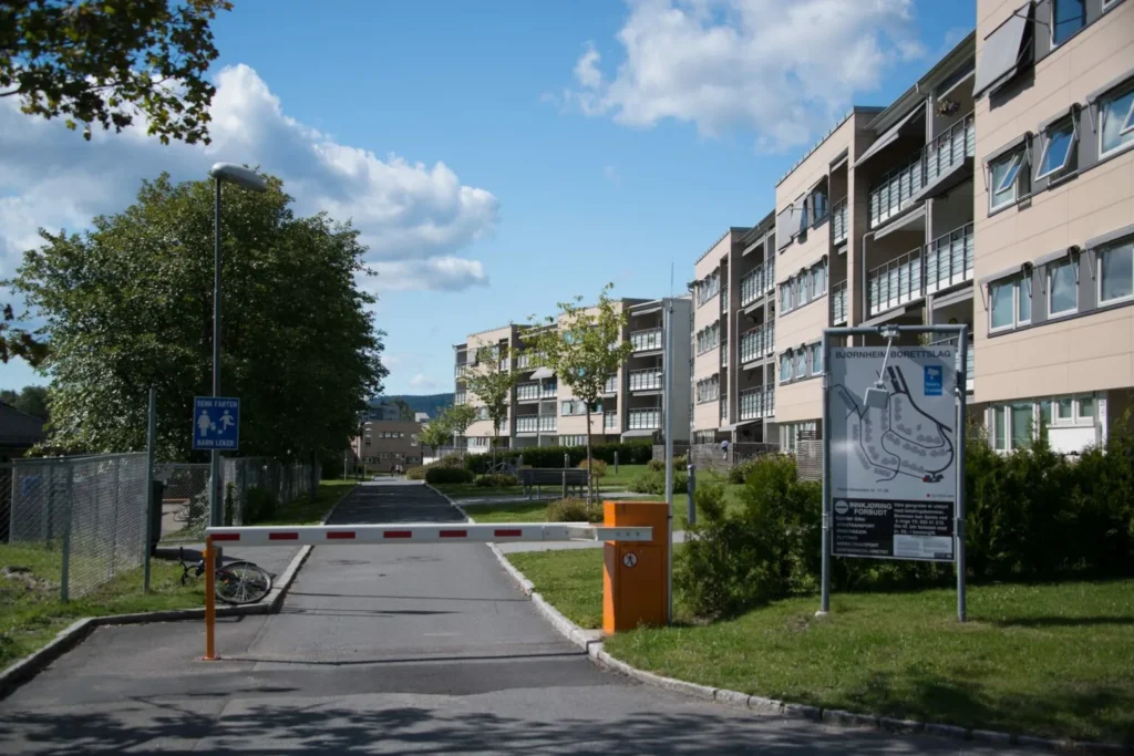 I det rolige boligområdet Bjørnheim Borettslag reiser beige bygårder seg under en klarblå himmel. En vei er forsiktig hindret av en senket barriere ved siden av en oransje kontrollboks. I nærheten hviler en sykkel ved gjerdet, med trær omtenksomt langs fortauet.