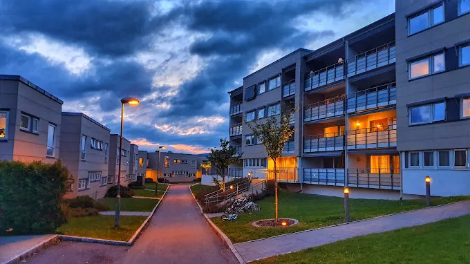 I skumringen viser de moderne bygårdene til Bjørnheim Borettslag opplyste vinduer og gatelykter. Himmelen, delvis overskyet med et snev av solnedgang, skaper et rolig bakteppe for stier og gressområder nedenfor. Beboerne har trygghet takket være Serwents ekspert rørfornying tjenester som sikrer kvalitetsinfrastruktur.