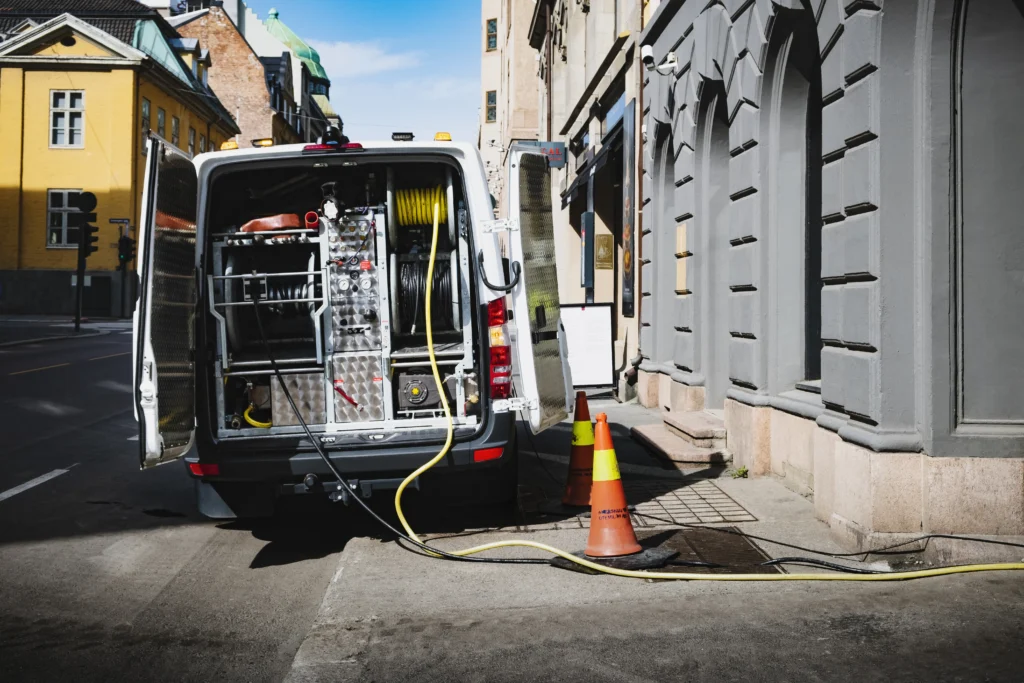 En bruksbil med åpne bakdører er parkert ved siden av en bygning i en bygate. Innvendig er utstyr, slanger og kabler for rørspyling synlige. To oransje trafikkkjegler er plassert nær et kum, med høytrykksslanger som strekker seg inn i den for effektivt vedlikehold av tette rør.