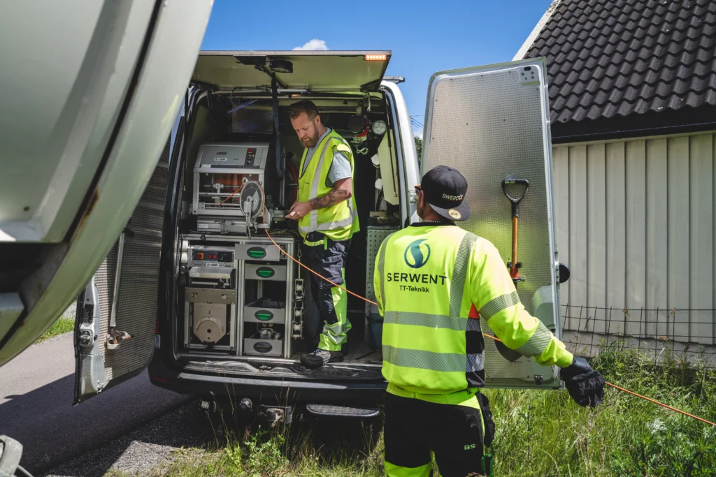 To bruksarbeidere i synlige klær driver rørinspeksjon, betjener utstyr fra åpen varebil utenfor et borettslag. Den ene står inne i varebilen og styrer en kabel, mens den andre observerer. Varebilen viser ulike verktøy og utstyr som er avgjørende for deres oppgave.