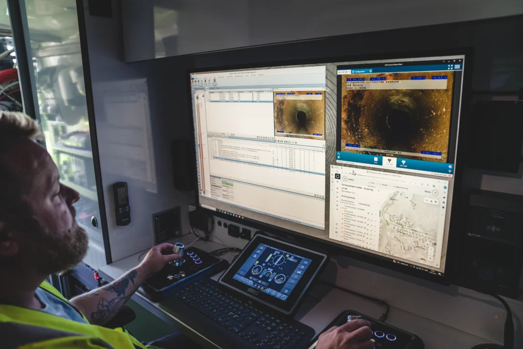 En person driver en kontrollstasjon med tre skjermer som viser rørinspeksjonsbilder, data og kart. Ved hjelp av kontroller og et tastatur analyserer de muligens underjordiske forhold mens de sjekker rør med kamera.