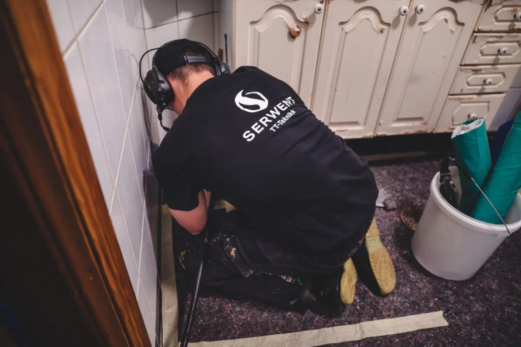 En person iført svart T-skjorte merket «SERWENT Technik» ligger på kne på gulvet, engasjert i rørfornying for Arbeidernes Bygge-Compagni III. De jobber på en del av veggen i et rom med hvite skap, ved hjelp av verktøy og beskyttende hodetelefoner. I nærheten ligger en bøtte og sammenrullet matte.