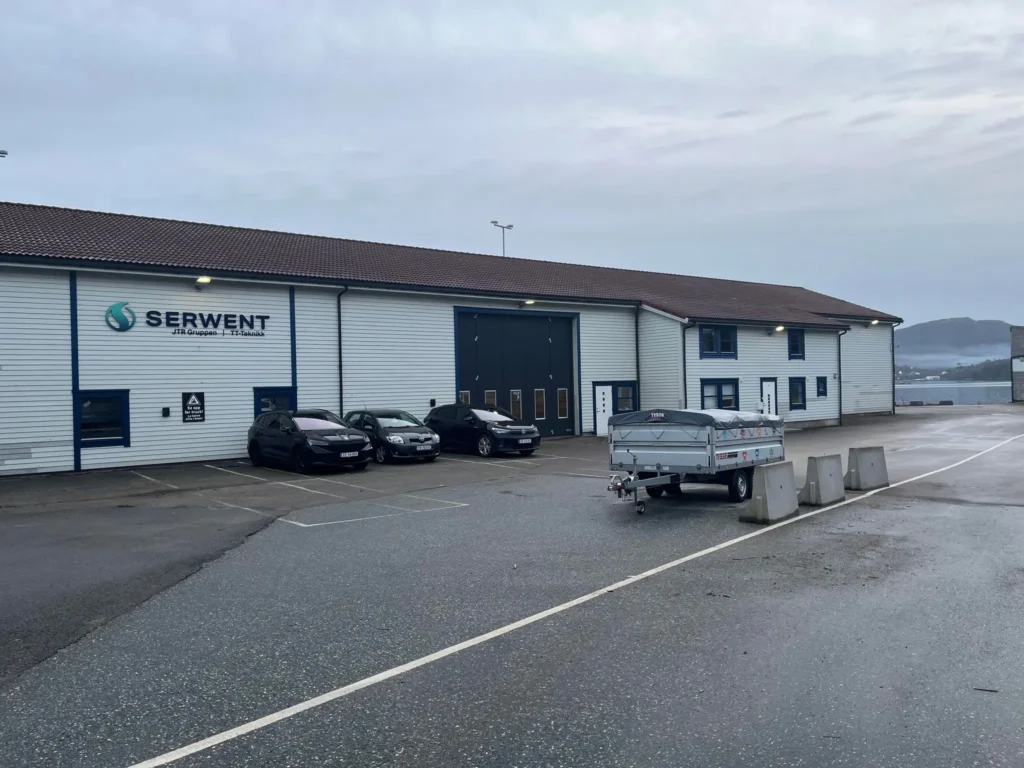 Et industrilager med "Serwent"-skiltet dominerer scenen, som stolt kan skryte av sitt ISO-sertifikat. En trailer og flere biler står parkert på det våte fortauet under en overskyet himmel, mens åsene strekker seg i det fjerne.