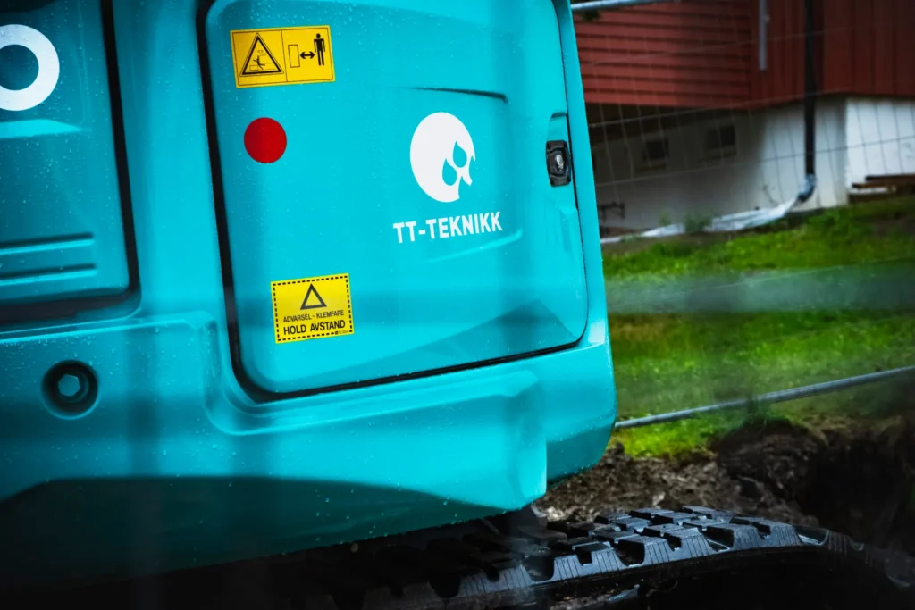 Nærbilde av et blågrønt anleggskjøretøy med logoen "TT-Teknikk", ofte sett på oljetankeranlegg, prydet med forsiktighetsklistremerker. Bakgrunnen avslører et gressområde og en uskarp struktur.