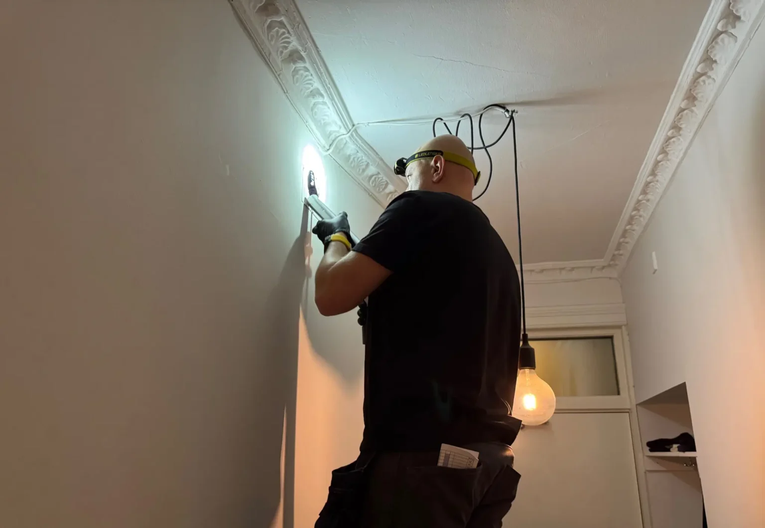 En person som har på seg hansker og hette er kyndig å installere eller reparere en lysarmatur eller ledninger i et hvitt tak i et borettslag. De står på en stige i et rom med utsmykket kronelist, mens to pendellamper henger grasiøst i bakgrunnen.