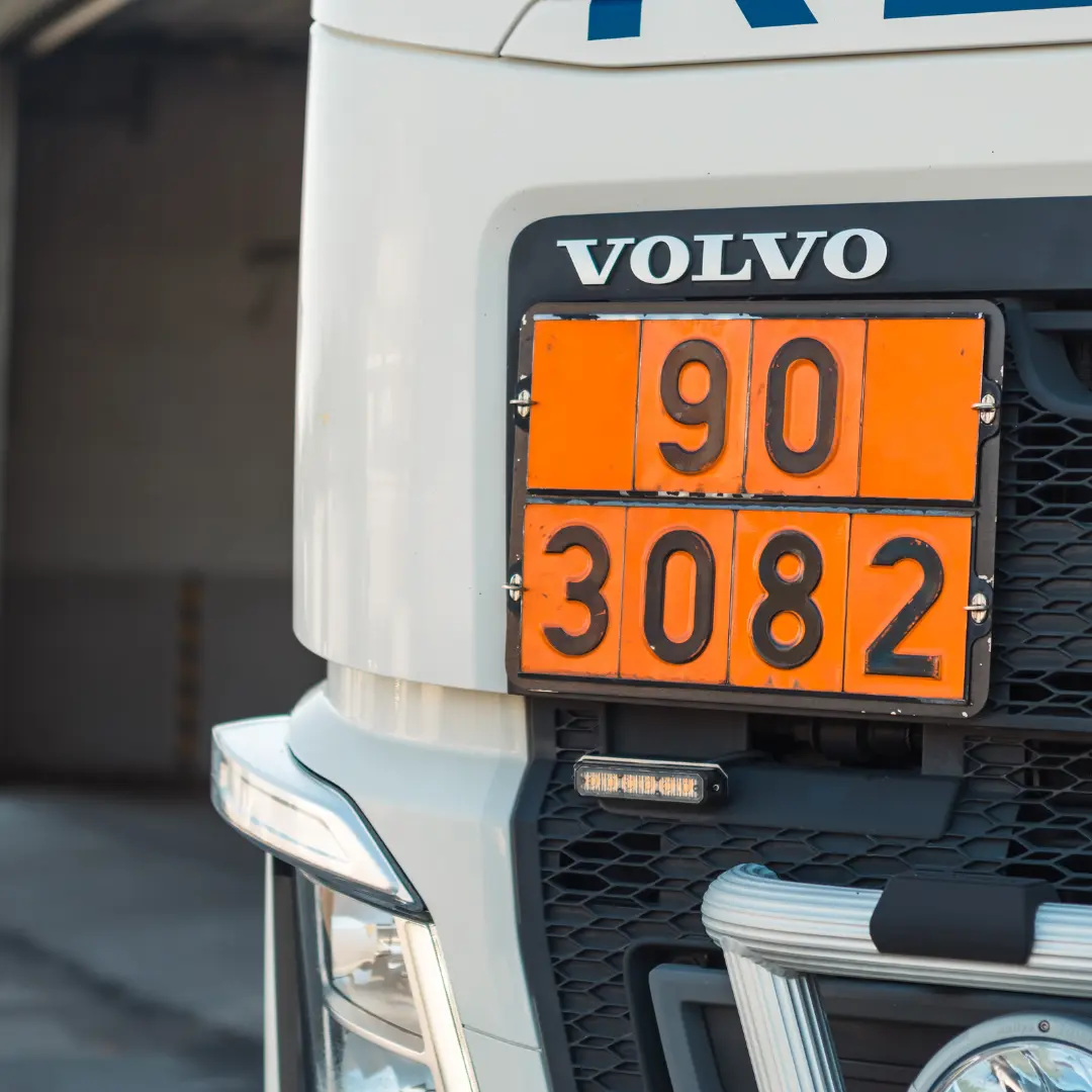 Närbild av fronten på en Volvo-lastbil som visar en orange faroskylt med siffrorna 90 och 3082. En del av lastbilens galler och strålkastare är synliga, med en suddig bakgrund av ett lager.