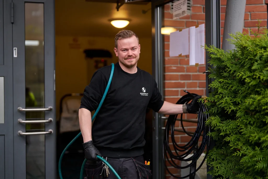 En person i svart genser og hansker, som ligner en ekspert på borettslag og sameier, står ved den åpne døren til en murbygning. De holder en opprullet kabel og blå slange, og ser til siden med et smil.