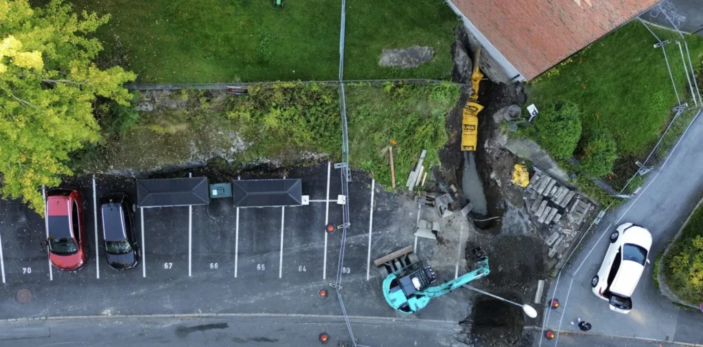 Flyfoto av et vellykket anleggsprosjekt med parkeringsplass ved siden av et bygg under oppføring. Flere biler er parkert på nummererte plasser. Anleggsmaskiner og materialer er synlige i nærheten av bygningen, ved siden av en delvis gravd grøft. Trær grenser til området på den ene siden.
