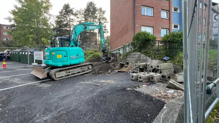 Gravemaskin En turkis gravemaskin står parkert på en byggeplass ved siden av en murbygning, som symboliserer et vellykket anleggsprosjekt. Hauger med skitt og betongblokker ligger spredt rundt, med trær og en parkeringsplass med søppelcontainere synlig i bakgrunnen.