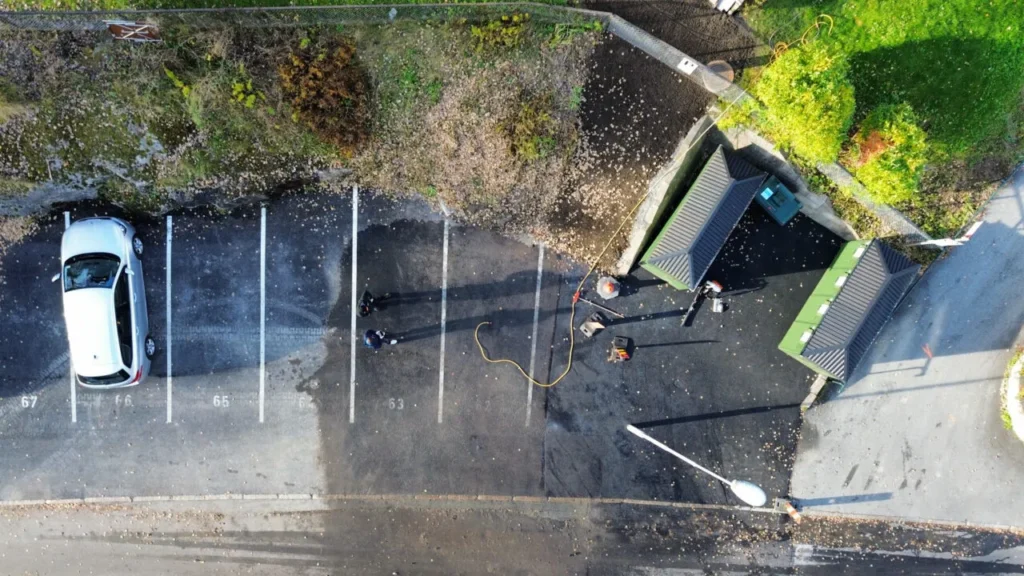Luftfoto av en parkeringsplass med seks plasser, fire okkupert av høytrykksvaskere og en slange. På plass 57 står en bil parkert. To små grønne bygninger øverst til høyre antyder et vellykket anleggsprosjekt, omkranset av gress og sti. Fallne løv er spredt rundt.
