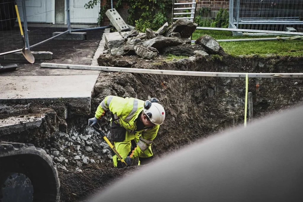 En flittig bygningsarbeider i hvit hjelme og gult sikkerhetsutstyr graver dyktig en grøft på den travle tomten, omgitt av maskineri, steiner og sikkerhetsbarrierer. Hans presisjon gjenspeiler de høye standardene for Krantjänster-praksis som er observert her.