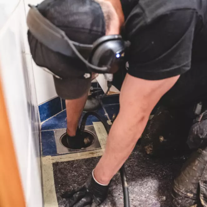 En person med hodetelefoner og hansker er fokusert på avloppsrensning i et flislagt område. Kledd i svart med en hette på, bruker de et verktøy for å nå inn i dreneringshullet, og jobber iherdig med å tømme det.