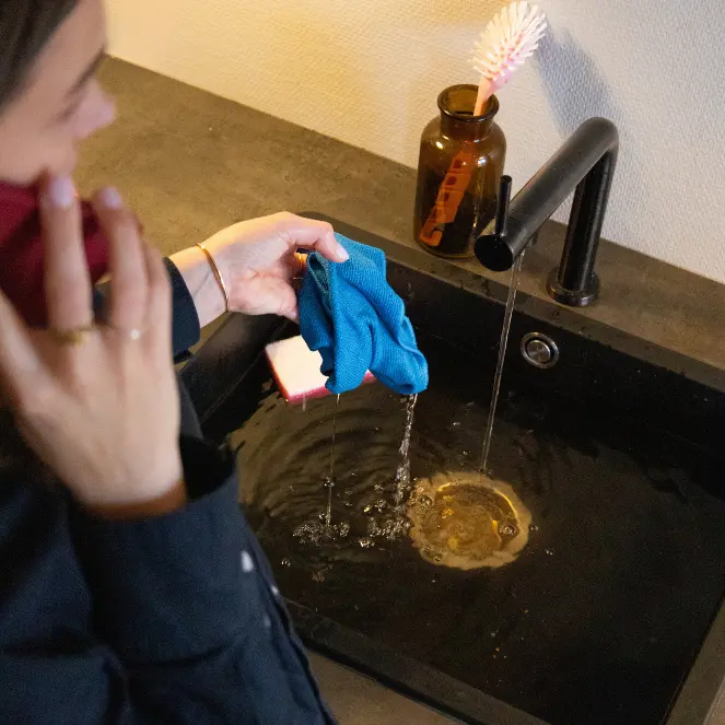 En person rengjør flittig en svart vask med en blå klut, og lar vannet strømme fra den elegante, svarte kranen. Engasjert i avloppsrensning holder de en rød gjenstand, mens en skrubbebørste hviler i en brun flaske like ved. Et mørkt erme antyder deres fokuserte oppgave.