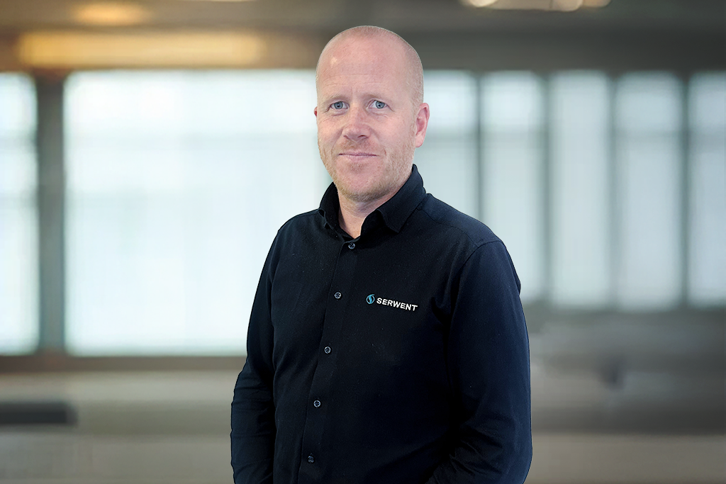 En mann med barbert hode og svart skjorte med SERWENT-logo står innendørs foran store vinduer og smiler lett til kameraet.