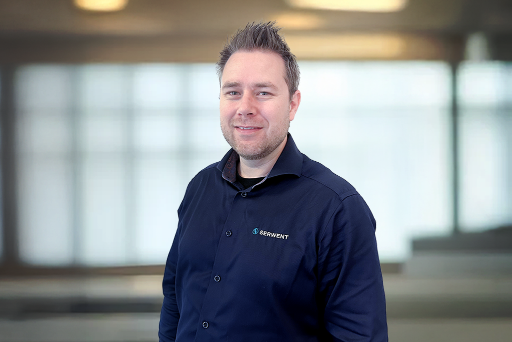 En mann med kort, spisshåret hår, iført en mørkeblå skjorte med SERVNEXT-logo, smiler til kameraet. Bakgrunnen er et kontor med store vinduer som er ute av fokus.