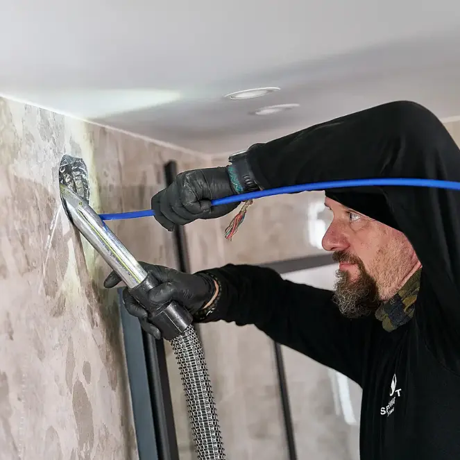 En person i arbeidsantrekk og hansker trer en blå kabel gjennom et hull i en mønstret vegg, nesten som om han orkestrerte en teknisk dans. De virker fokuserte, og bruker verktøy for å hjelpe i prosessen midt i det som virker som et innendørsrom under bygging, som ligner et bassengfyllningsprosjekt.