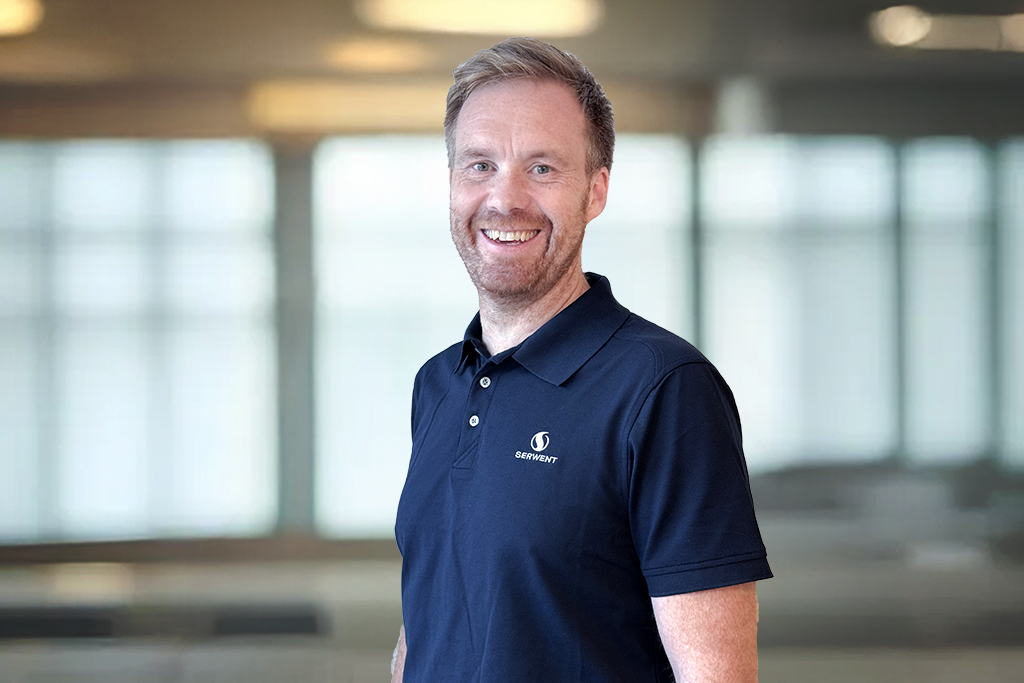 En smilende mann med lysebrunt hår og skjegg, iført marineblå poloskjorte med firmalogo, står i et moderne, lyst kontorlokale med store vinduer i bakgrunnen.