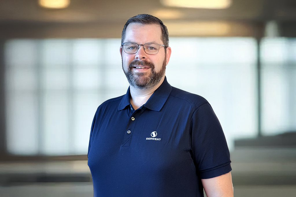 En mann med kort, brunt hår, skjegg og briller smiler mens han står innendørs. Han har på seg en marineblå poloskjorte med en firmalogo der det står Servment. Bakgrunnen er et uskarpt kontorlandskap.