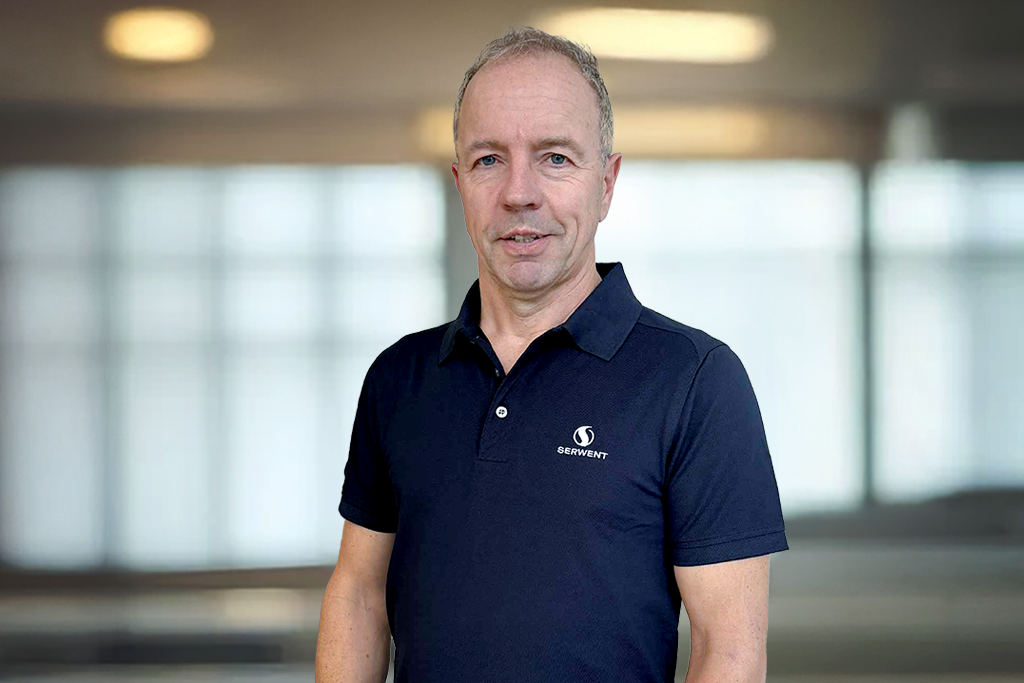 En middelaldrende mann med kort, grått hår og mørkeblå poloskjorte står innendørs foran store, uskarpe vinduer. Han ser inn i kameraet og smiler lett.