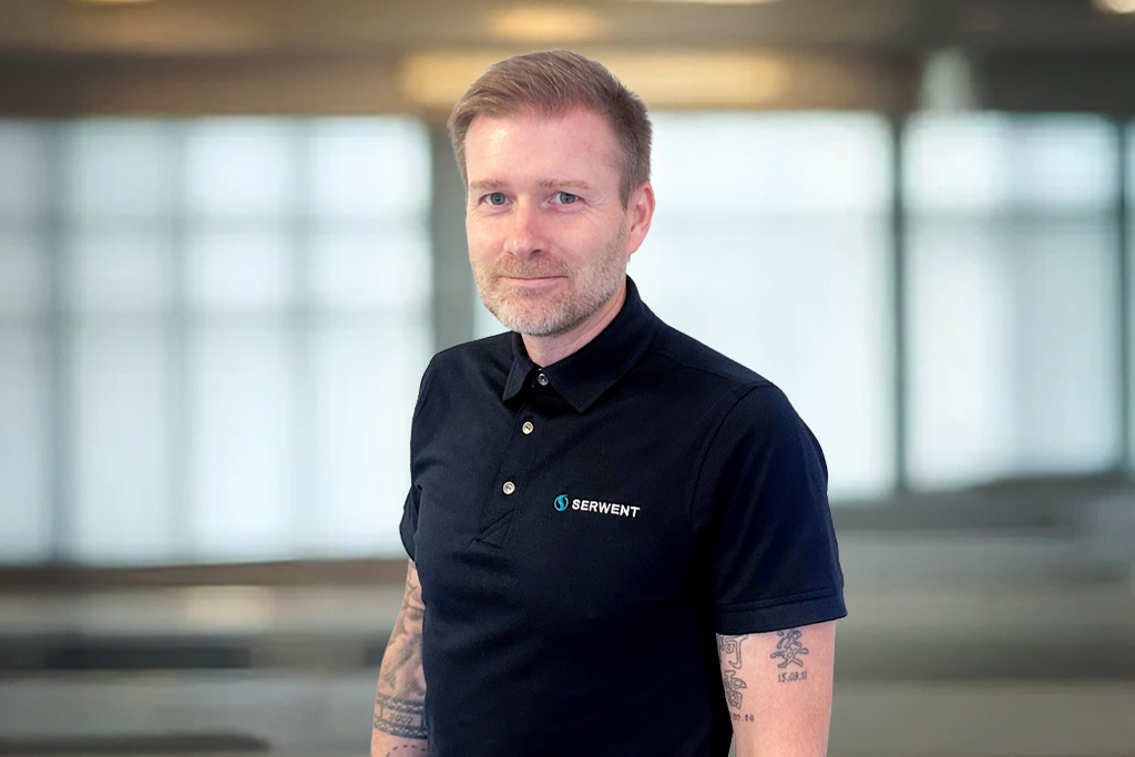 En mann med kort, lyst hår og tatoveringer på armene står innendørs, iført marineblå poloskjorte med Serivent-logo. Bakgrunnen er uskarp og viser store vinduer og et mykt, naturlig lys.