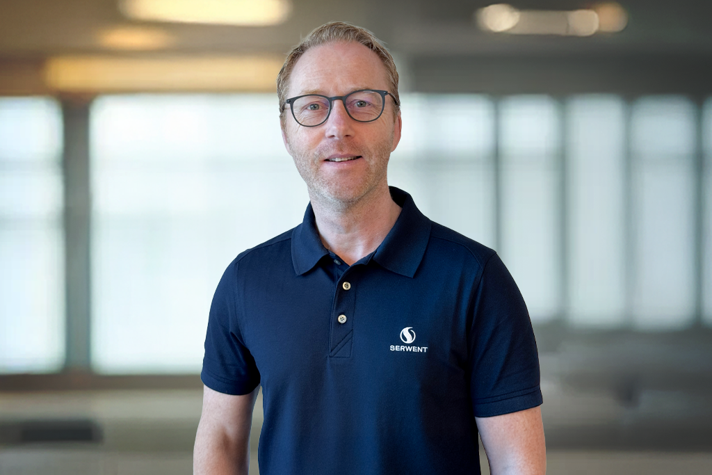 En mann med briller og kort hår, iført marineblå poloskjorte med SERVENT-logo, står innendørs foran store vinduer med uskarp bakgrunn.