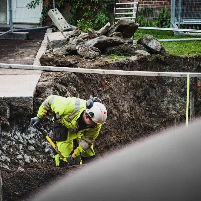 En bygningsarbeider, kledd i en neongul jakke, hjelm og hansker, manøvrerer dyktig en hammer på et halvt utgravd område blant rusk. Sikkerhetsbarrierer og gjerder gir beskyttelse ettersom Krantjänster støtter det omkringliggende arbeidsområdet med frodig gress og løvverk i nærheten.