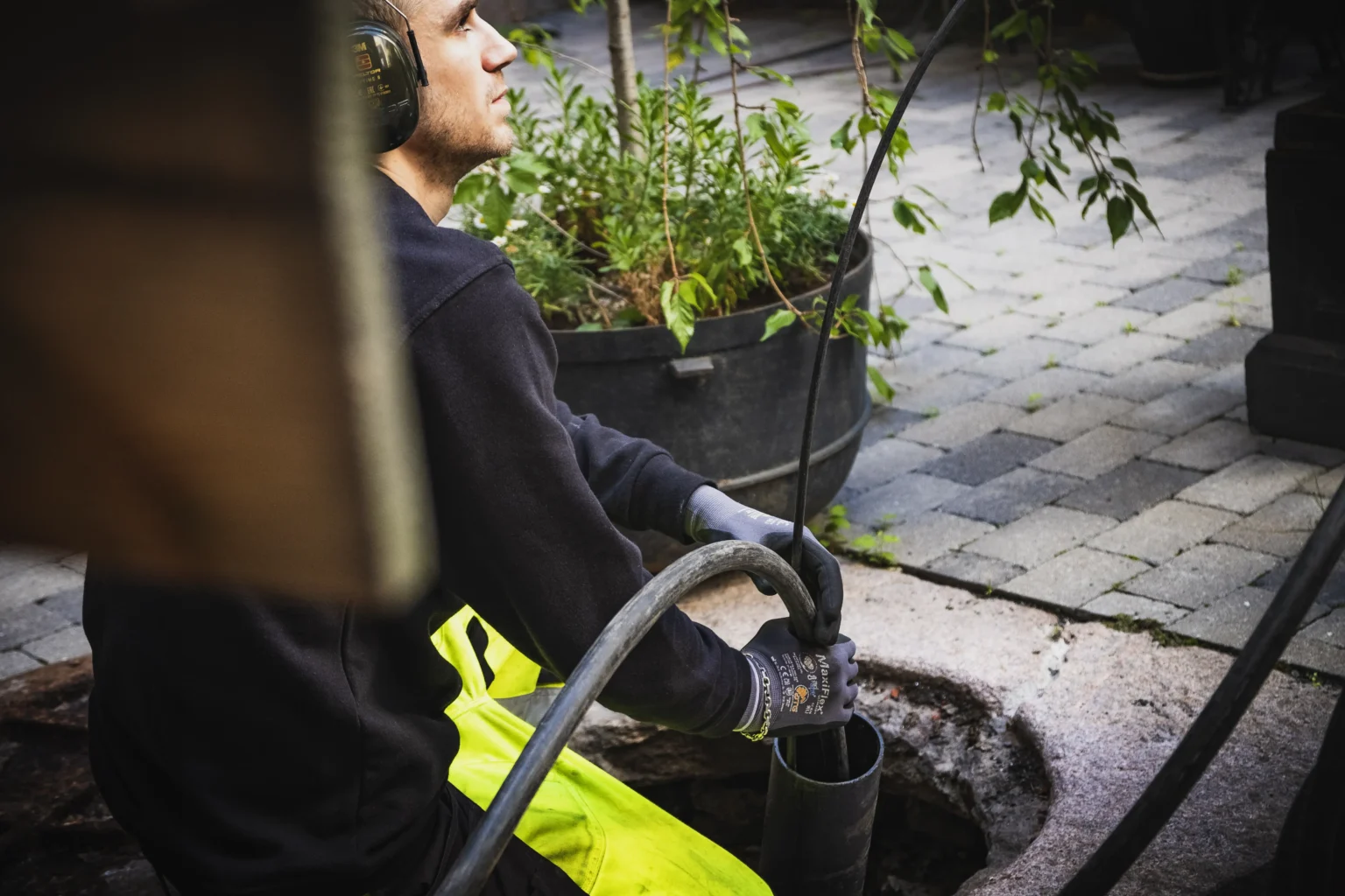 En arbeider iført verneutstyr og hansker bruker en slange til å utføre vedlikehold eller rengjøring på et utendørs avløp eller kum, med planter og belegningsstein synlig i bakgrunnen – et typisk trinn i rørfornyingsprosessen.
