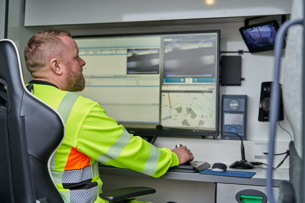 En person som har på seg en synlig jakke, bruker datautstyr med flere skjermer. Skjermene viser kart og feeder for overvåkingskameraer. Innstillingen ser ut til å være et kontrollrom eller operasjonssentral.