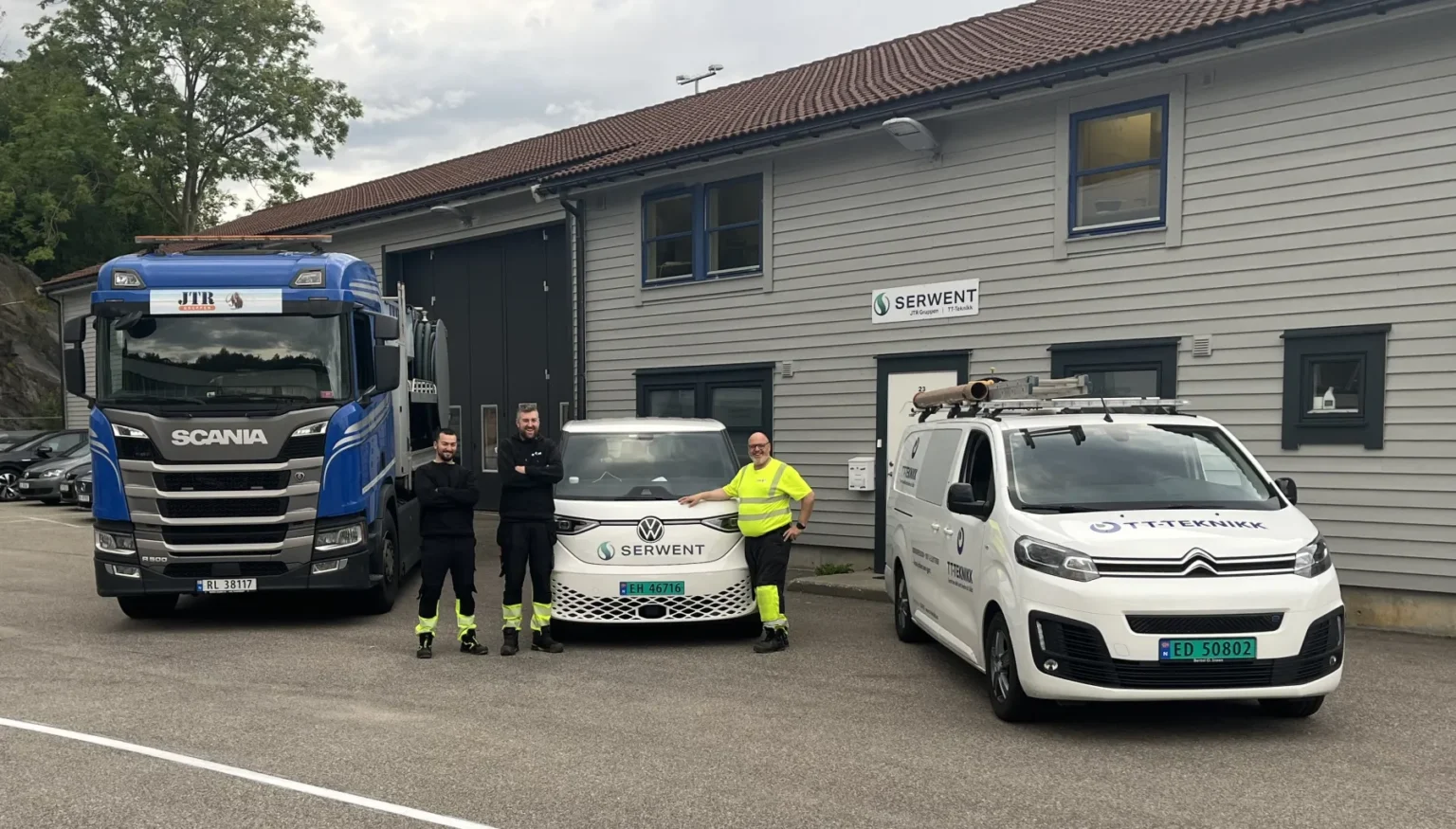 Tre personer står foran en blå lastebil, en hvit varebil og en hvit bil merket med "Serwent" nær en grå bygning. Det er skiltet «Serwent» og «Trefikk» på bygget. Området er oversiktlig med trær i bakgrunnen.