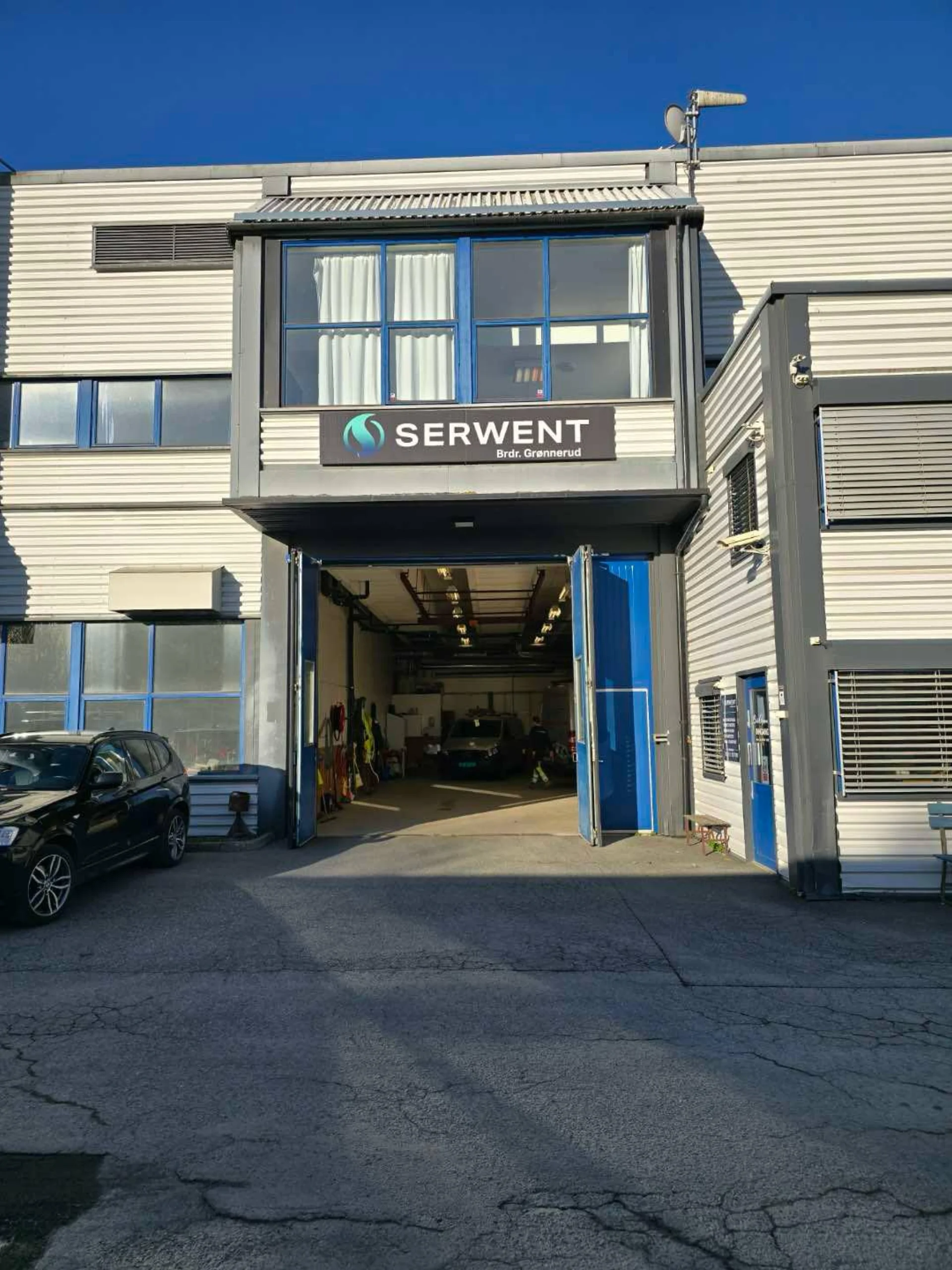 En toetasjes industribygning med blå og grå detaljer, store åpne garasjeporter og et skilt med teksten SERWENT over inngangen. En svart bil står parkert til venstre, og sollyset kaster skygger på bakken.