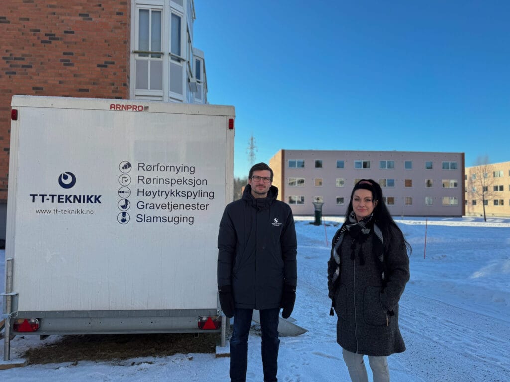 To personer står foran en TT-Teknikk servicebil i en snødekt gate. Varebilen har firmalogoer og tekst på norsk. Begge personene har på seg vinterklær. Det er bolighus og en klar blå himmel i bakgrunnen.