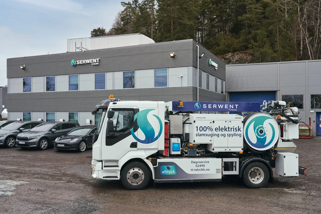 Elektrisk avfallsbil parkert foran et moderne industribygg med "Serwent"-skilt. Lastebilen er hvit med grønn og blå grafikk, med tekst "100% elektrisk". Flere biler er parkert i nærheten. Trær og overskyet himmel er i bakgrunnen.