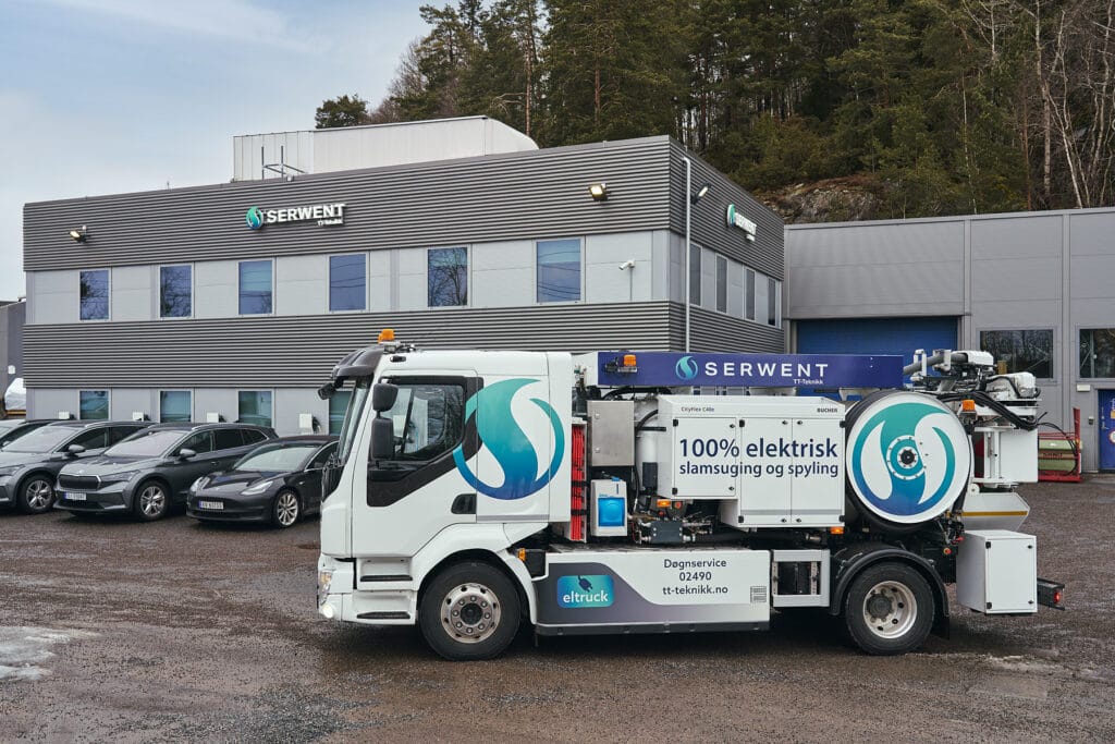 En elektrisk servicebil med firmamerke står parkert foran et moderne industribygg merket «Serwent». Flere biler står parkert ved siden av lastebilen, og trær er synlige i bakgrunnen.